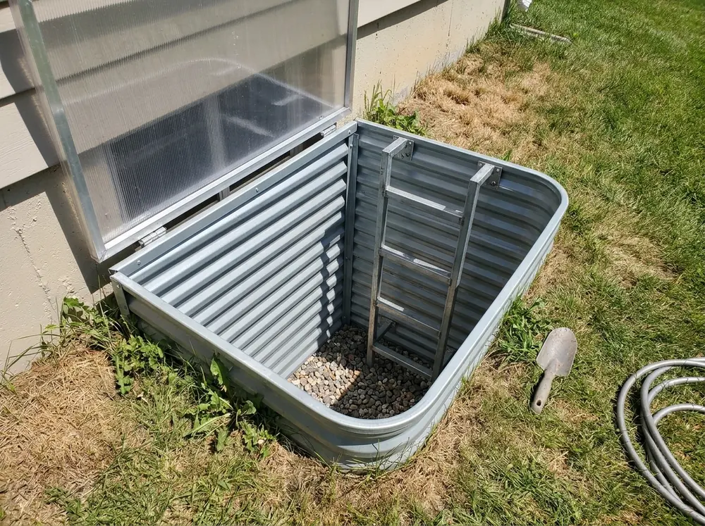 A gray corrugated metal egress window well with a fixed steel ladder and a clear hinged cover over the top